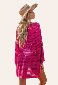 Różowa szydełkowana narzutka o luźnym kroju, z diamentowym wzorem, głębokim dekoltem i szerokimi rękawami, w zestawie z brązowym słomkowym kapeluszem.