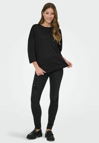 Joven mujer sonriendo, vestida con una blusa negra de manga larga holgada, jeans skinny negros rasgados y mocasines negros, de pie sobre un fondo liso.