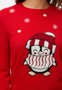 Sarkans adīts džemperis, kurā attēlots pingvīns ar svītrainu sarkano un baltu cepuri un šalli. Augšdaļā ir sniegpārsliņu raksti.