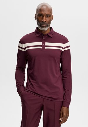 Hombre con barba gris que lleva una camisa polo de manga larga burdeos con dos rayas horizontales blancas en el pecho y pantalones a juego.