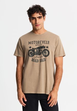 Młody mężczyzna z kręconymi włosami, ubrany w beżowy t-shirt z czarną grafiką motocykla i tekstem "Motorcycle Races American Road Race."