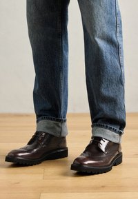 Zapatos brogue de cuero marrón con un acabado pulido, que presentan una suela negra robusta, cordones y perforaciones decorativas. Combinados con jeans de mezclilla doblados.