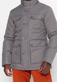 Giacca grigia imbottita con colletto alto, due tasche sul petto e dettagli con bottoni. Presenta un tessuto texturizzato e una silhouette dritta.