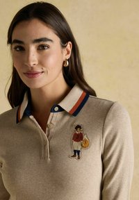 Frau mit langen braunen Haaren, die ein beiges Poloshirt mit navyblauen und rostfarbenen gestreiften Kragen sowie einem gestickten Fuchs-Patch auf der Brust trägt.
