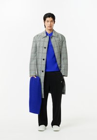 Grå rutigt trenchcoat över en blå tröja och svarta byxor. Modellen håller en blå stickad scarf. Skorna är vita och tyget ser slätt ut.