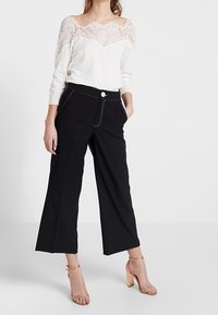 Maglione bianco con scollatura in pizzo, pantaloni neri a gamba larga con tasche frontali e tacchi nudi con cinturini, caratterizzati da cuciture a contrasto.