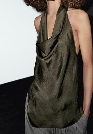 Femme portant un haut sans manches en satin vert olive drapé avec un décolleté profond et un pantalon gris, sur un fond noir et blanc.