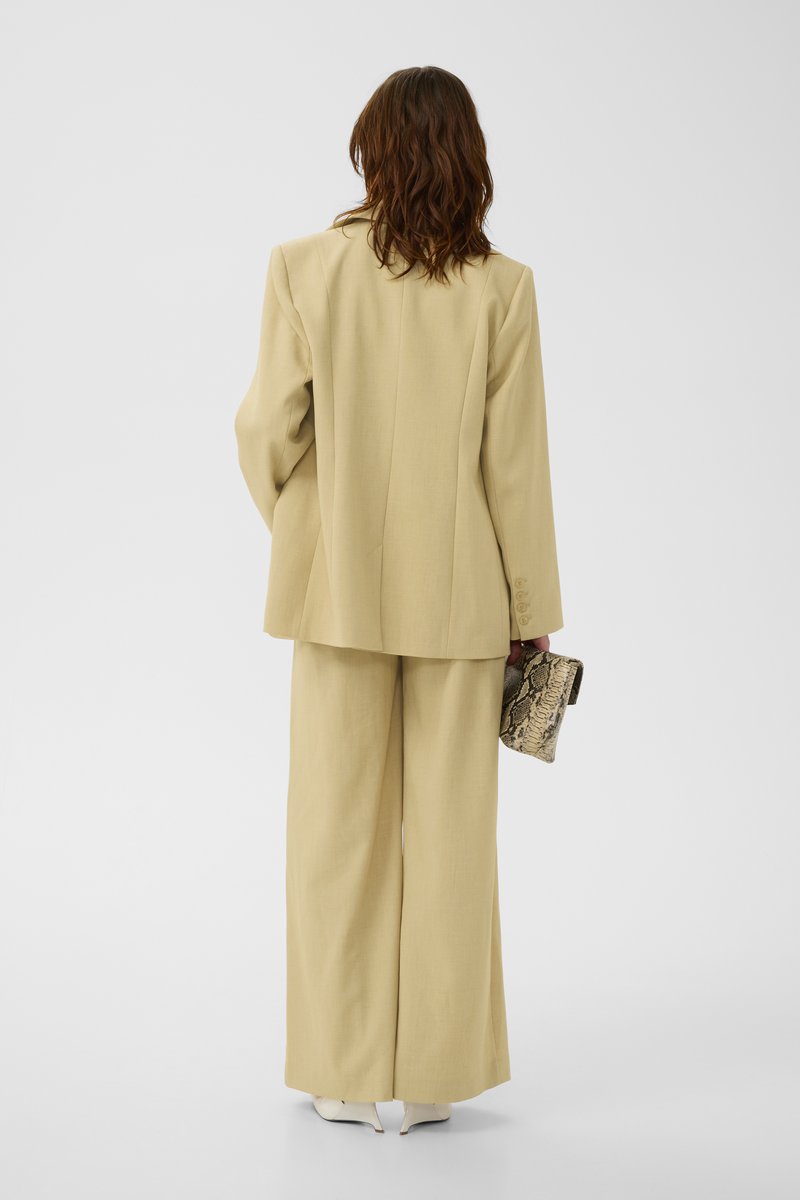 Femme aux cheveux bruns mi-longs portant un costume beige oversize et tenant un petit sac à main à motif peau de serpent, debout de dos.