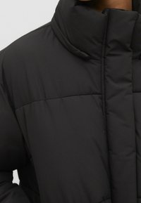 Sort pufferjakke med høj krave, quiltet design og glat tekstur, med skjulte lukninger og afslappet pasform.