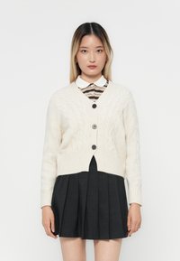 Cárdigan de punto trenzado color crema con cuello en V, tres botones y dobladillo acanalado, combinado con una camisa de cuello a rayas y una falda plisada oscura.