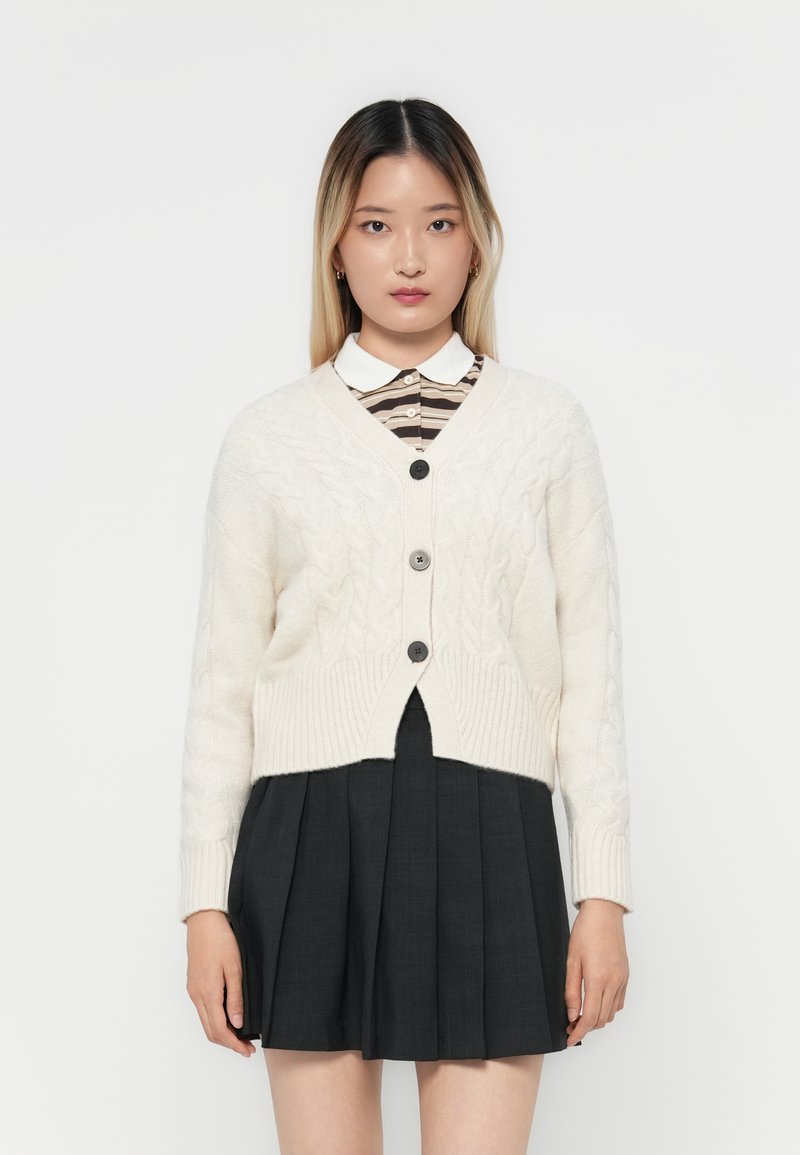 Cardigan beige a trecce con scollatura a V, tre bottoni e fondo a costine, abbinato a una camicia a righe con colletto e una gonna plissettata scura.