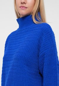 Pull en grosse maille bleu col roulé avec un motif texturé et un col côtelé. Matériau doux, coupe décontractée et lignes horizontales distinctes.