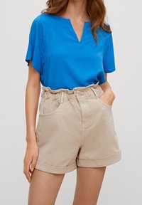 Kobieta w jasnoniebieskiej bluzce z krótkim rękawem i beżowych szortach z wysokim stanem, z podwiniętymi mankietami, z jedną ręką w kieszeni, widoczny tali.