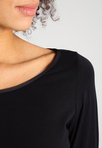 Top nero con una texture liscia, scollo ampio e un sottile dettaglio rifinito sulla spalla. Presenta un primo piano del tessuto e del design.