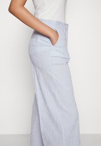 Personne portant un haut blanc à manches courtes et un pantalon large à rayures bleu clair, debout de côté avec une main dans la poche.