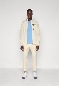 Beige jakke med glidelås og logo, lys blå t-skjorte, beige bukser med avsmalnende mansjetter, og hvite sneakers med mørke detaljer.