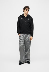 Νεαρός άνδρας που φοράει μαύρο hoodie με φερμουάρ και το λογότυπο BMW Motorsport, λαμπερό ασημί παντελόνι, μαύρα παπούτσια και στρογγυλά μαύρα γυαλιά ηλίου, στέκεται με τα χέρια στις τσέπες.