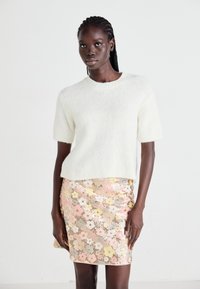 Hvit, kortermet, fluffy genser parret med et tettsittende rosa blomstrete paljett-miniskjørt med gule og hvite blomster på en teksturert bakgrunn.