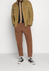 Pantalon en velours côtelé marron, chemise vert olive, pull noir avec bande beige, baskets noires, chaussettes blanches, coupe décontractée avec poches avant.