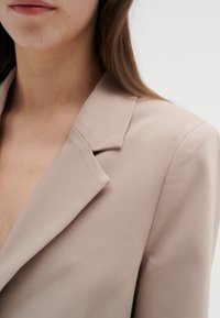 Beige blazer med slagkrage, slät textur och skräddarsydd passform. Närbild visar detalj av slaget och enkla designelement.