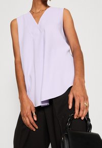 Vaatetuslaventeli ilman hihnoja, jossa on V-kaula-aukko, laskeutuu pehmeästi vartalon yli, korostaen teksturoitua, väljästä istuvuutta, yhdistetty tummiin teksturoituihin housuihin.