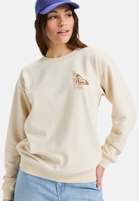 Beige sweatshirt met ronde hals, gemaakt van zachte katoen, met een bruine tropische graphic en "ROXY" tekst op de linkerkant van de borst.