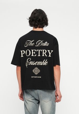 Osoba s krátkymi kučeravými vlasmi nosí čierne tričko s béžovým textom "The Delta Poetry Ensemble" a dekoratívnym symbolom, spárované s vyblednutými modrými džínami.