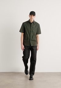 Groen gemusterd overhemd met korte mouwen, relaxte pasvorm, gecombineerd met donkerdenim broek. Een zwarte pet en schoenen maken de outfit compleet. Effen achtergrond.