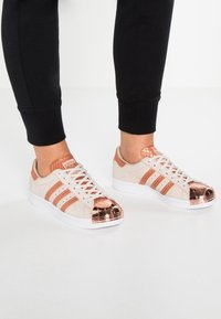 Beige suède sneakers met rosegouden metalen neuskappen, drie rosegouden strepen en witte rubberen zolen. De veters zijn wit en geweven.