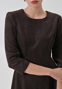 Dunkelbraunes Kleid aus strukturiertem Stoff mit einem Fischgrätenmuster, hat einen runden Ausschnitt und dreiviertellange Ärmel.