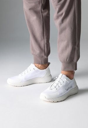 Personne portant des baskets blanches de sport avec des semelles texturées et un pantalon de jogging gris sur un fond neutre.