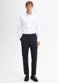 Man in een strakke witte blouse met lange mouwen, zwarte cargobroek en zwarte instap schoenen staat met zijn handen in de zakken.