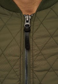 Gestepptes olivgrünes Jacke mit Reißverschluss, hochgestelltem Kragen und schwarzem Reißverschlussanhänger. Strukturierter Stoff mit Rautenmuster.