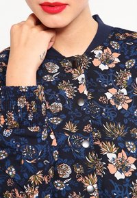 Blommig bomberjacka i marinblått tyg, med olika blommönster i rosa, vitt och grönt. Knappar och elastiska ärmslut ger extra detalj.