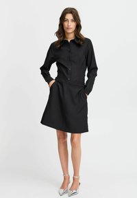 Schwarzes Kleid mit Knopfverschluss an der Vorderseite, langen Ärmeln und tailliertem Schnitt. Hergestellt aus glattem Stoff, mit einem schwingenden Rock und praktischen Seitentaschen.