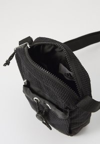 Bolsa negra texturizada con cierre de cremallera, correa ajustable y un bolsillo frontal con un cordón. Interior revestido con tela suave, con la marca visible.