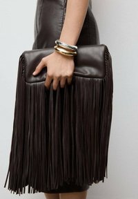 Tamno smeđi kožnati neseser s dugačkim resama, pravokutnog oblika i glatke teksture. Metalne narukvice ukrašavaju ruku koja drži torbu.