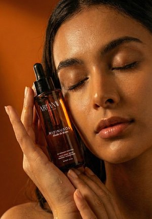 Flacon compte-gouttes en verre ambré étiqueté "Rizinusöl Huile de Ricin", tenu près du visage d'une personne à la peau lisse et saine.