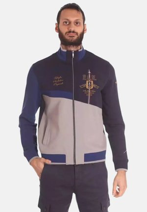 Giacca zip-up blu navy e grigia con colletto alto, dotata di tasche frontali, accenti ricamati e design a blocchi di colore contrastanti.