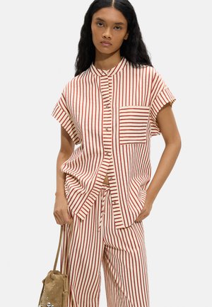 Kobieta w kremowej i czerwonej koszuli w pionowe paski z krótkim rękawem zapinanej na guziki oraz z pasującymi spodniami, trzymająca beżową torbę z troczkiem i ćwiekami.