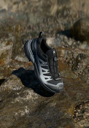 Scarpa da trekking Salomon nera e grigia con sistema di allacciatura rapida poggiata su una superficie rocciosa esterna ruvida.