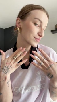 Γυναίκα που δείχνει περιποιημένα νύχια, φοράει πολλά δαχτυλίδια και χρυσές σκουλαρίκια, με ορατά τατουάζ στα χέρια και τα μπράτσα, ντυμένη με ένα ανοιχτόχρωμο πουκάμισο.