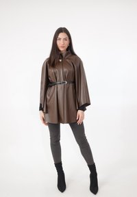 Bruine cape van namaakleer met een hoge kraag, ceintuur in de taille. Gecombineerd met grijze skinny jeans en zwarte enkellaarsjes in sokstijl.