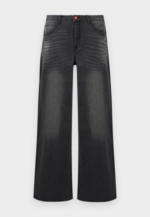 Persoon draagt wijd uitlopende vervaagde zwarte jeans, gecombineerd met wit-zwarte sneakers, een zwart jack en een donkergrijze top.