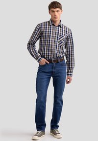 Camisa de manga larga a cuadros en azul marino, verde y blanco; jeans ajustados de color azul oscuro; cinturón marrón; zapatillas grises; pose de pie contra un fondo neutro.