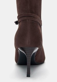 Stivale alto in suede marrone con tacco nero texturizzato, con una fascia attorno alla caviglia e una finitura liscia e lucida.