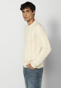 Suéter de punto trenzado color crema con cuello redondo y puños acanalados, combinado con jeans azules. La textura presenta patrones tejidos elevados.
