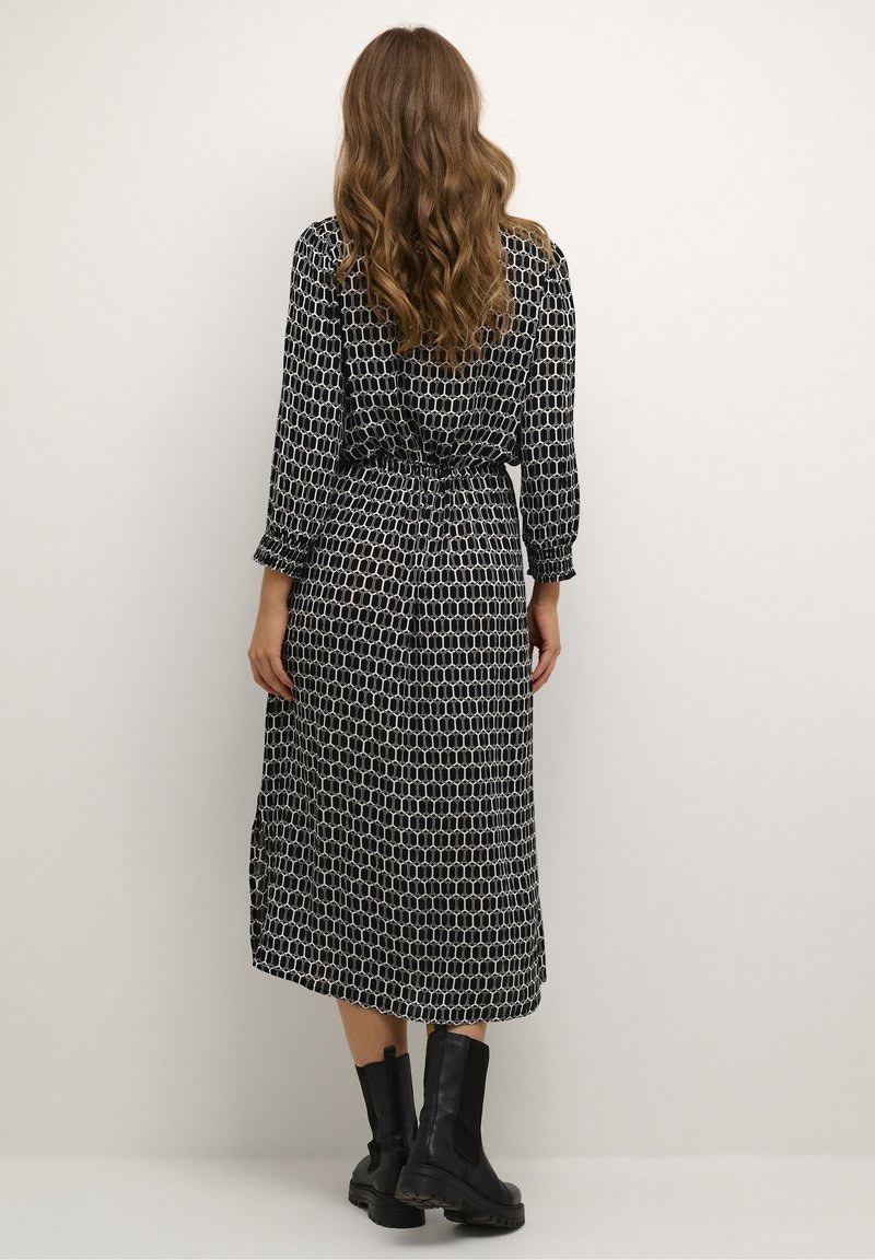 Femme aux longs cheveux ondulés portant une robe midi noire et blanche à motifs et des bottines noires, debout face à un mur clair uni.