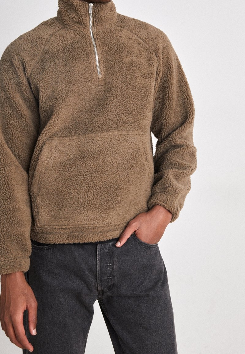 Brauner Fleecepullover mit hohem Kragen, Reißverschlussdetail und Fronttasche. Strukturierter Stoff mit weichem Finish, kombiniert mit dunkler Jeans.