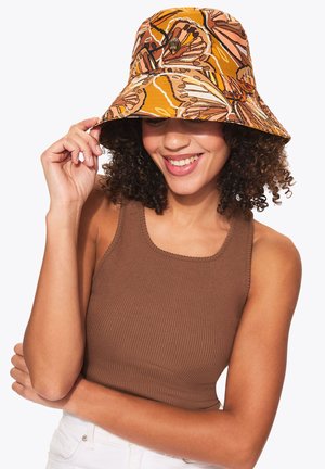 Donna sorridente con capelli ricci che indossa una canottiera marrone a coste e un cappello a secchiello arancione con stampa di farfalle, tenendo la tesa del cappello con una mano.